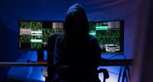 Ένας ανώνυμος χάκερ με κουκούλα μπροστά από υπολογιστή σε σκοτεινό δωμάτιο